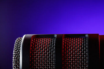 A microphone sits on a table with a dark background. The mesh part reflects red and purple light. The scene shows a recording session setup. Equipment is ready for use.