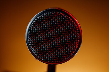 A microphone sits in focus with a metal mesh top. The background is brown with some red light. This setup often appears in recording sessions or live performances at night.