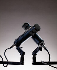 Two microphones rest on a stand in a music studio, prepared for recording. Cables connect them as they await the artist to start. A neutral background surrounds them.