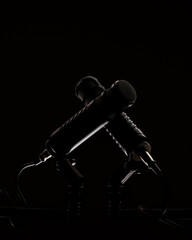 Two microphones are positioned in a crossed manner on a dark surface. The background is black, creating a focus on the microphones. Cables extend from their bases, indicating they are ready for use.
