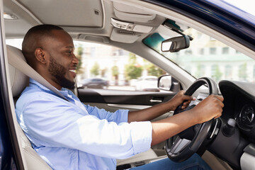 Side view profile portrait of joyful African American man driving car, going to work or on trip during summer vacation. Happy guy riding his brand new nice car, doing test drive in dealership showroom © Prostock-studio