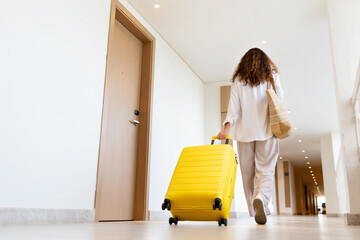 Adult caucasian woman walking away in hotel hallway with suitcase and shoulder bag, calm solo...