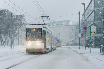 Winter city street tram heavy snowfall shows public transport operating icy condition reduced visibility. Snow covered tracks urban buildings mobility challenge safety risk harsh seasonal weather