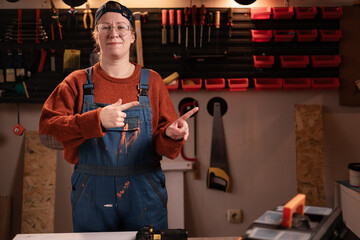 Woman joiner wears workwear in workshop scene with tools hanging on wall point index fingers aside on space for text indicate. female carpenter late evening in authentic garage.