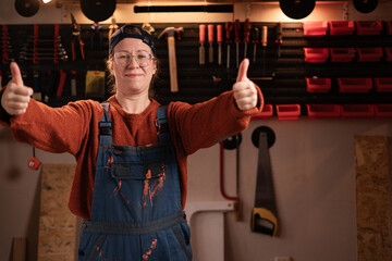 Woman joiner wears workwear in workshop scene with tools hanging on wall shows thumbs up stretching arms to camera. female carpenter late evening in authentic garage.