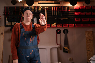 Woman joiner wears workwear in workshop scene with tools hanging on wall making stop gesture...