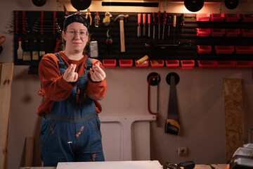 Woman joiner wears workwear in workshop scene with tools hanging on wall making money gesture....