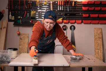 Female Worker applying white paint on wooden board in carpentry workshop. Wood finishing and painting process