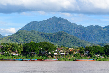 Fototapeta premium A vibrant riverside scene in Luang Prabang, Laos, with dense tropical trees, scattered houses, and long wooden boats lining the Mekong river. The backdrop features forested hills and a blue sky dotted