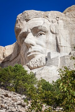 Close view of Abraham Lincoln carved in granite at Mount Rushmore National Memorial, framed by evergreens under a clear blue sky in the Black Hills of South Dakota.