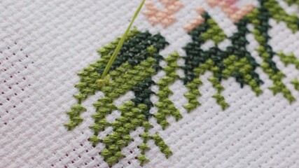 Extreme close up embroiderer guiding green thread through needle into cross stitch leaves on white canvas, blue nail visible, precise hand movement, fine weave texture, calm outdoor daylight mood