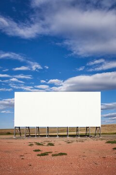 A large white drive in movie screen stands on the prairie beneath a wide blue sky with soft clouds in rural South Dakota, offering ample copy space and a clean, minimal scene.