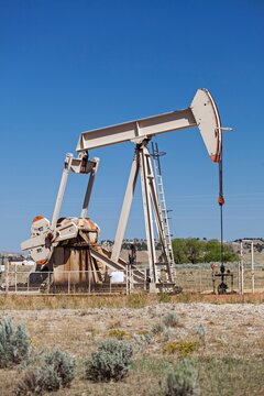 An oil pump jack operates in Rozet, Wyoming, set against a clear blue sky. The industrial structure highlights the region's energy production and resource extraction industry.