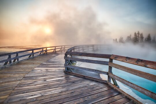 Steaming hot spring at sunrise with wooden boardwalk curving through West Thumb Geyser Basin in Yellowstone National Park.