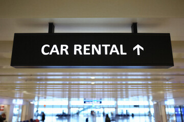 Car rental sign with white text on black background in airport terminal with people walking around in background
