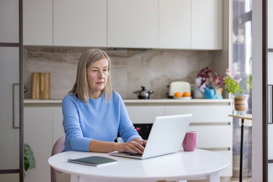 Senior woman using a laptop for remote work or online learning, typing on the keyboard while sitting at a kitchen table, embracing technology in her home environment - Powered by Adobe