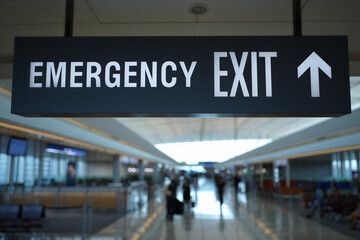 Emergency exit sign with white arrow pointing up in airport terminal hallway with people walking in background