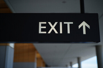 Exit sign with white arrow pointing upwards on black rectangular board in a building corridor with blurred background
