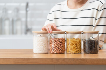 Young Asian woman organizing various healthy ingredients like grains, nuts, and pasta in clear glass jars, focusing on a nutritious longevity lifestyle and kitchen organization.