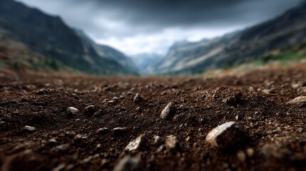 Close-up view of textured soil with scattered stones, showcasing the rich earth tones and natural elements, framed by a dramatic mountainous landscape under a cloudy sky