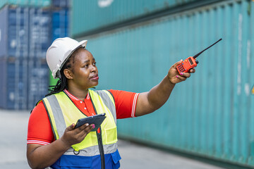African female supervisor in a safety vest holding a tablet and using a walkie-talkie.