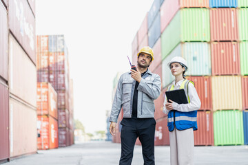 Professional male and female logistics supervisors wearing safety uniforms and hard hats, using a walkie-talkie and a digital tablet while working together at a maritime shipping container.