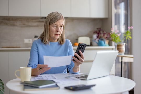 Senior woman at kitchen table reviewing household bills and paperwork, using laptop and smartphone for online banking, budgeting and retirement planning with focused concentration