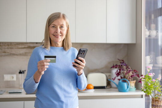 Senior woman in a modern kitchen smiling while holding a smartphone and debit card, making secure mobile banking or online shopping transactions at home with ease and confidence