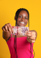 A smiling young woman with braided hair holds a 200 Ghanaian Cedi note toward the camera. She wears a pink top against a vibrant yellow background, her expression joyful and engaging