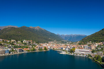 Fototapeta premium High-angle panorama of Omegna on Lake Orta in northern Italy, showing modern waterfront buildings, a densely built town center, and forested hills with rugged alpine peaks in the distance beneath a
