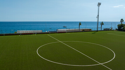 Aerial view of a modern artificial turf football pitch situated on the coast. Wide shot of the center circle of a soccer field. In background there is a calm deep blue sea and a clear horizon. © Stefano Tammaro