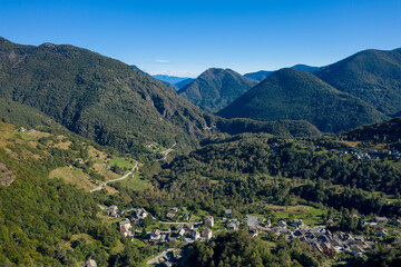 Fototapeta premium Aerial view of the small mountain village of Finero nestled in a lush, forested alpine valley surrounded by rolling hills and rugged peaks. The clear blue sky and vibrant greenery highlight the