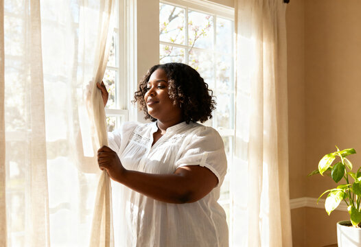 Black woman opening curtains to let in the bright morning sunlight at home. Hopeful person looking out the window for a new day and fresh start