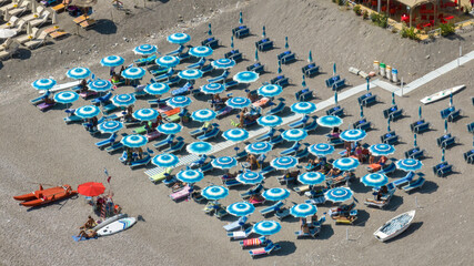 Aerial view of a beach club with blue and white striped umbrellas on grey pebbles. Features a red lifeguard station with a rescue boat and a white walkway for visitors. It is a summer morning in Italy