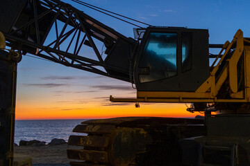 Construction crane silhouetted against dramatic ocean sunset on Portuguese coast at Furadouro
