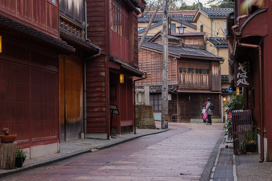 View of warm red-brown wooden buildings lining a winding cobblestone street, as a figure strolls through the historic district, Kanazawa, Ishikawa, Japan.
