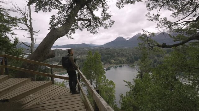 Male traveler with a backpack standing at a scenic wooden balcony overlooking the lush forests and mountains of Bahia Mansa in Villa La Angostura, Patagonia