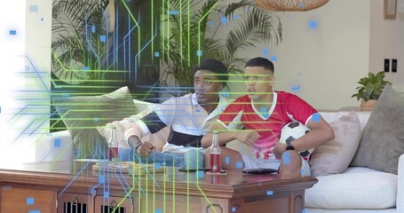 Watching two men in red jersey on sofa in living room, holding soccer ball, digital overlay