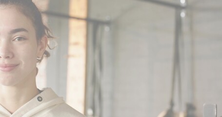 Smiling adult female wearing light hoodie standing at gym with rig, ropes, brick column, copy space