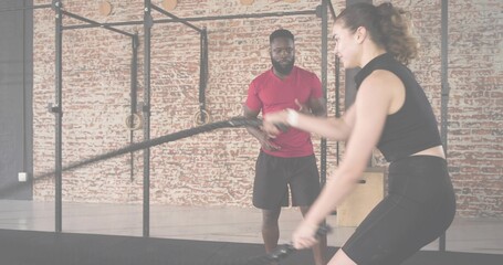 Swinging woman in black top and shorts doing battle ropes in studio, coach observing, copy space