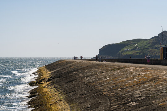 View of weathered stone pier stretches into the shimmering sea under a clear sky, people stroll along the pier towards the green headland, Howth, Dublin, Ireland.