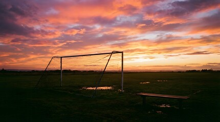 Atmospheric sports photography of silhouetted soccer goal at sunset with dramatic clouds, rain puddles reflections, empty wooden bench, and quiet rural football field