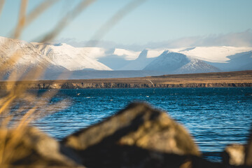 View of tranquil waters meeting the rugged coastline, leading to snow-capped mountains under a serene sky, Saudarkrokur, Skagafjordur, Iceland.