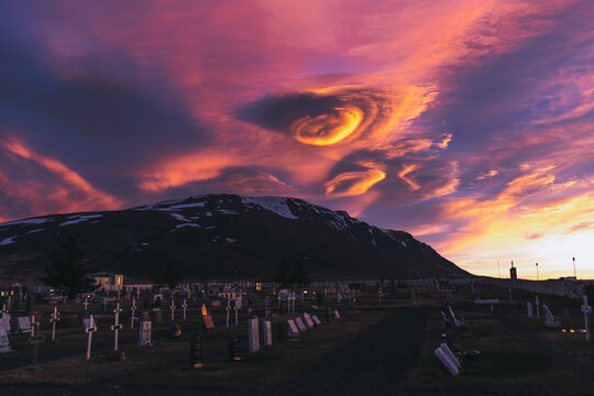 View of headstones dotting a tranquil graveyard beneath a dramatic sky painted with vivid hues of pink and orange against a backdrop of a snow-touched mountain, Saudarkrokur, Skagafjordur, Iceland.