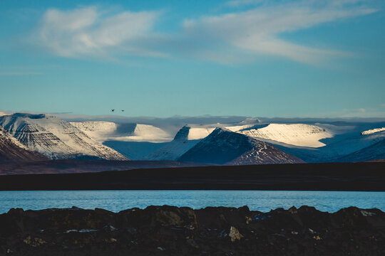 View of snow-capped mountains bask in the sunlight against a bright blue sky, separated by a dark band of land and water, Saudarkrokur, Skagafjordur, Iceland.