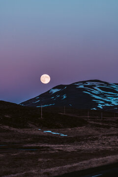 View of the full moon glows against a backdrop of dusky purple and blue skies over the shadowy hills in Saudarkrokur, Skagafjordur, Iceland.