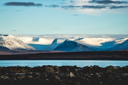 View of snow-capped mountains rise majestically beyond the tranquil waters, framed by rugged rocks under a serene sky, Saudarkrokur, Skagafjordur, Iceland.