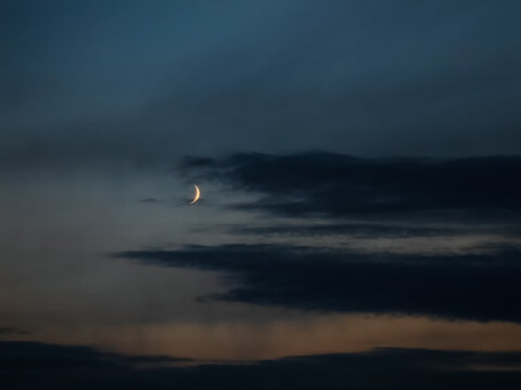 A waxing moon shines brightly in the dark evening sky against a backdrop of clouds after sunset, a new moon against a gray-blue background
