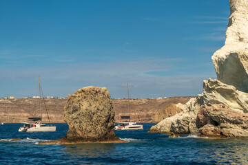 Sailboat passing rocky sea stack near coastal cliffs under blue sky