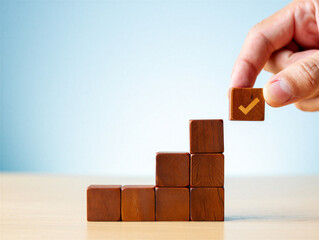 Hand placing wooden block with yellow checkmark on top of stacked blocks on table in front of blue background